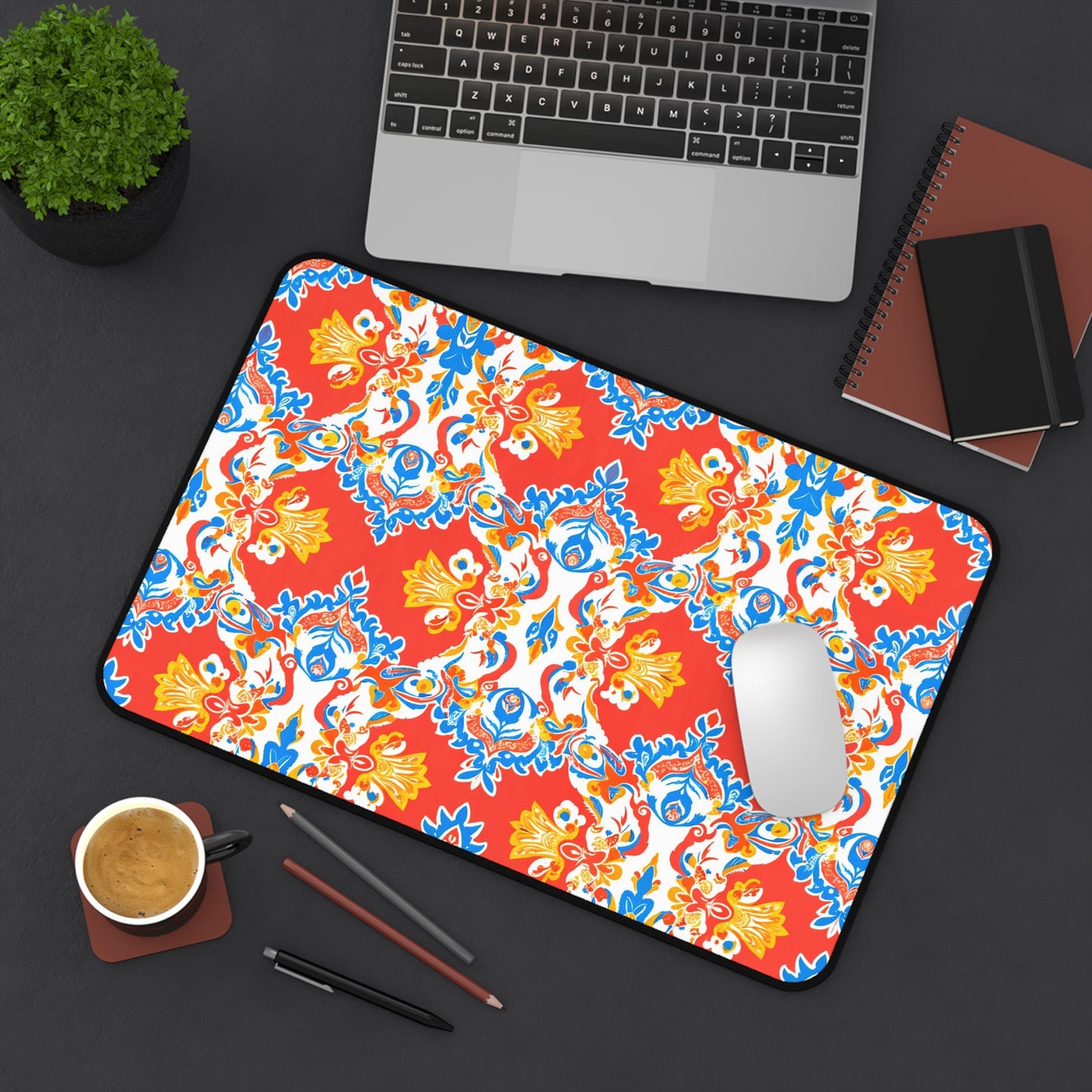 A large neoprene desk mat with a vibrant red, blue, yellow, and white floral pattern sits on a dark desk, alongside a silver laptop, brown notebook, black notepad, two pencils, a cup of coffee, and a small potted plant.