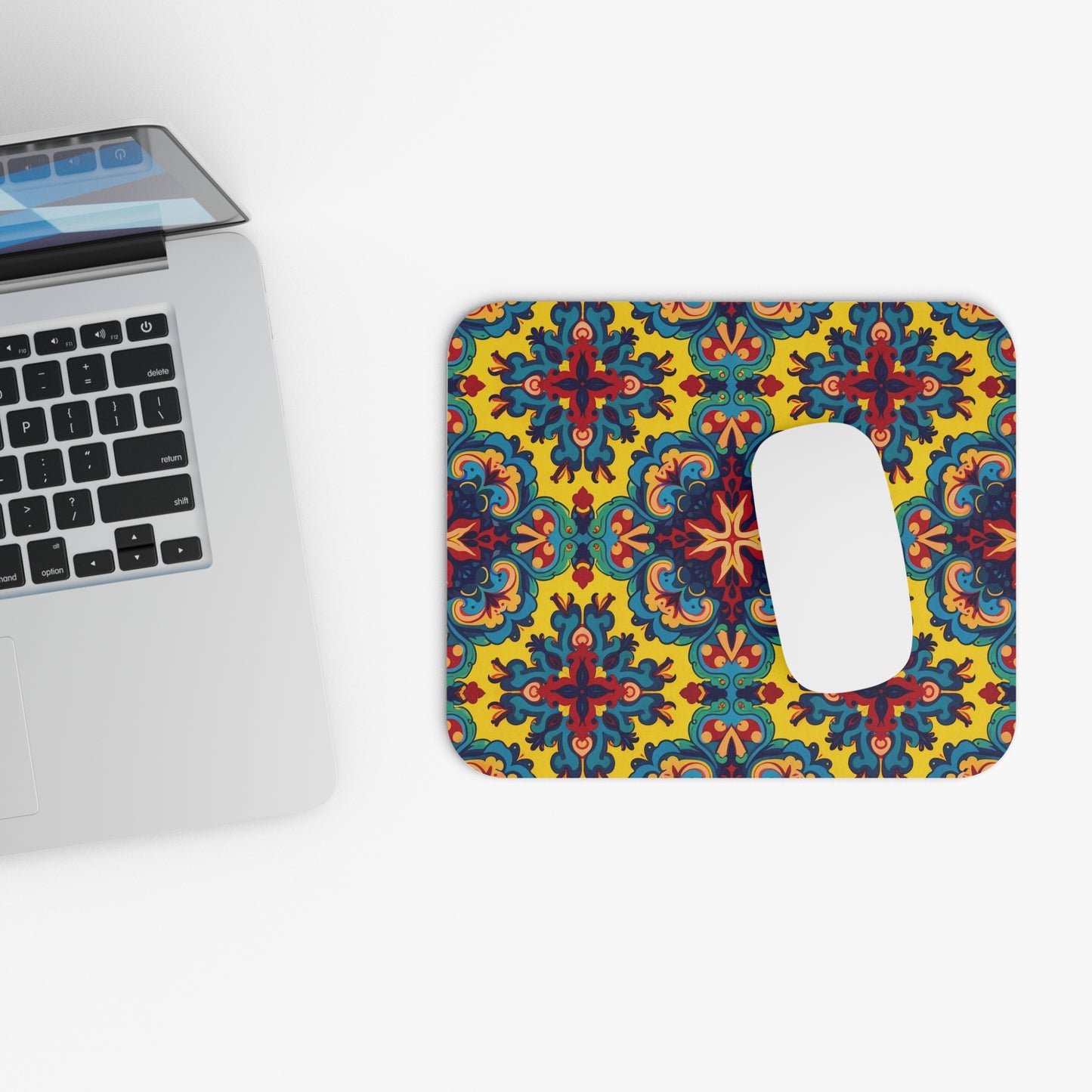 A silver laptop sits on a white surface next to an African-inspired mouse pad with a rectangular slot. The mouse pad showcases a vibrant, symmetrical floral pattern in blue, red, and green on a bright yellow background.
