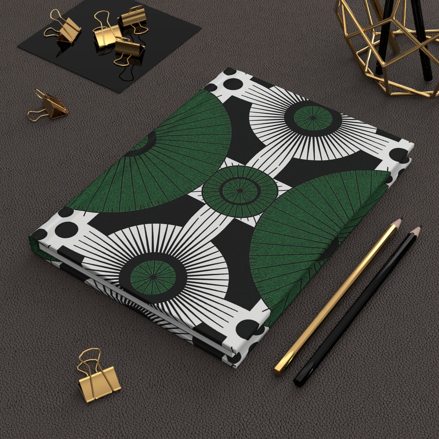 An African-inspired wellness journal with a bold black, white, and green geometric pattern lies on a textured brown desk. Nearby are a gold pen, a black pencil, a gold decor piece, and several gold binder clips on a black tray.