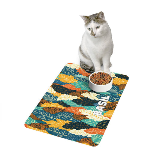 A white and gray cat sits beside a food bowl on an African-inspired, leaf-patterned mat. The mat features orange, yellow, teal, and blue leaves with the word BASIL printed in white letters. The background is plain white.
