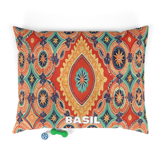 A rectangular, stylish pet bed pillow with a vibrant geometric and floral pattern in orange, blue, yellow, and teal. BASIL is printed in white at the bottom center. Below are a blue ball and a green chew toy, perfect for your pets.