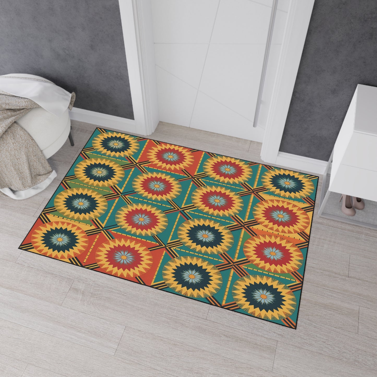 A colorful rectangular rug with African-inspired aesthetics and a repeating geometric starburst pattern in red, yellow, teal, and dark blue lies on light wooden floor in front of a white door. Grey walls and a bed with a beige blanket are at the left edge.