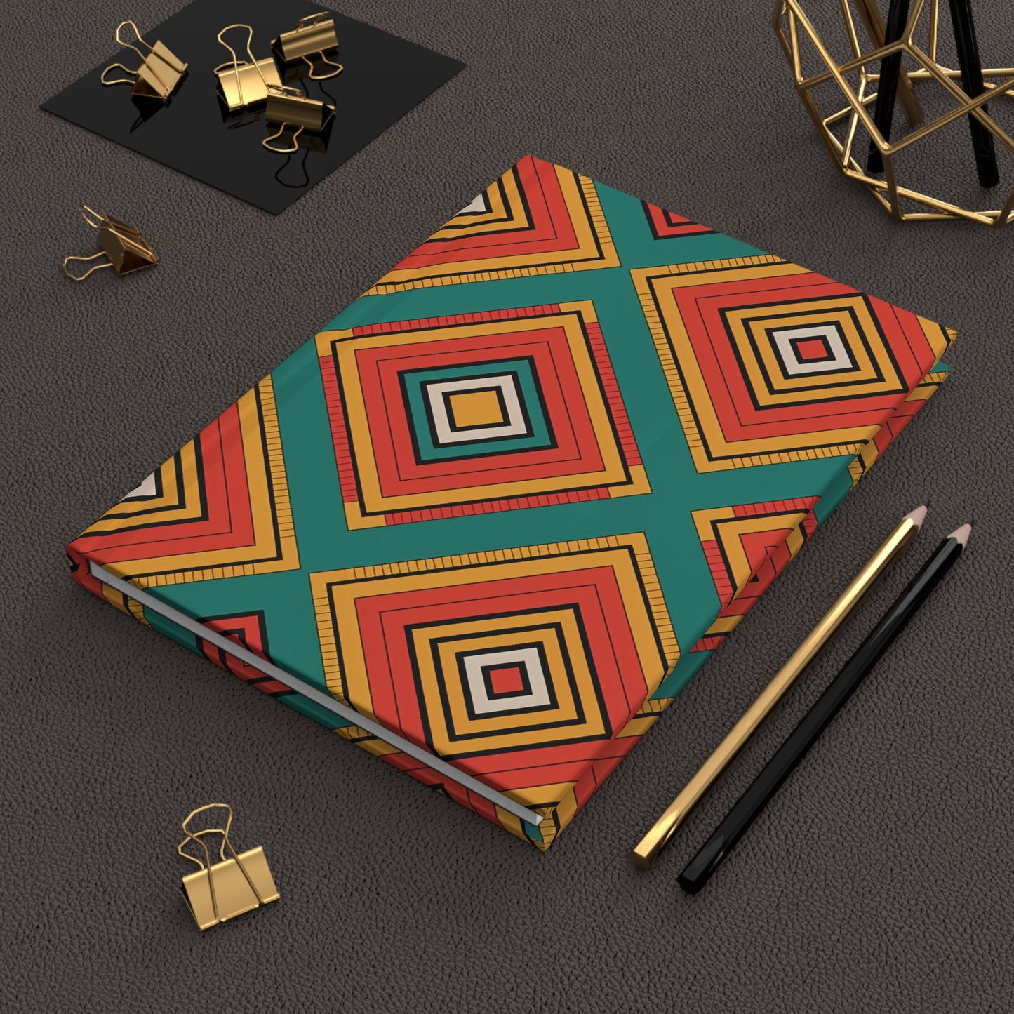 A geometric hardcover notebook with a bold diamond pattern in red, gold, teal, and black lies on a textured dark brown surface. Nearby are gold binder clips, a black and gold pencil, and a gold geometric decor piece.