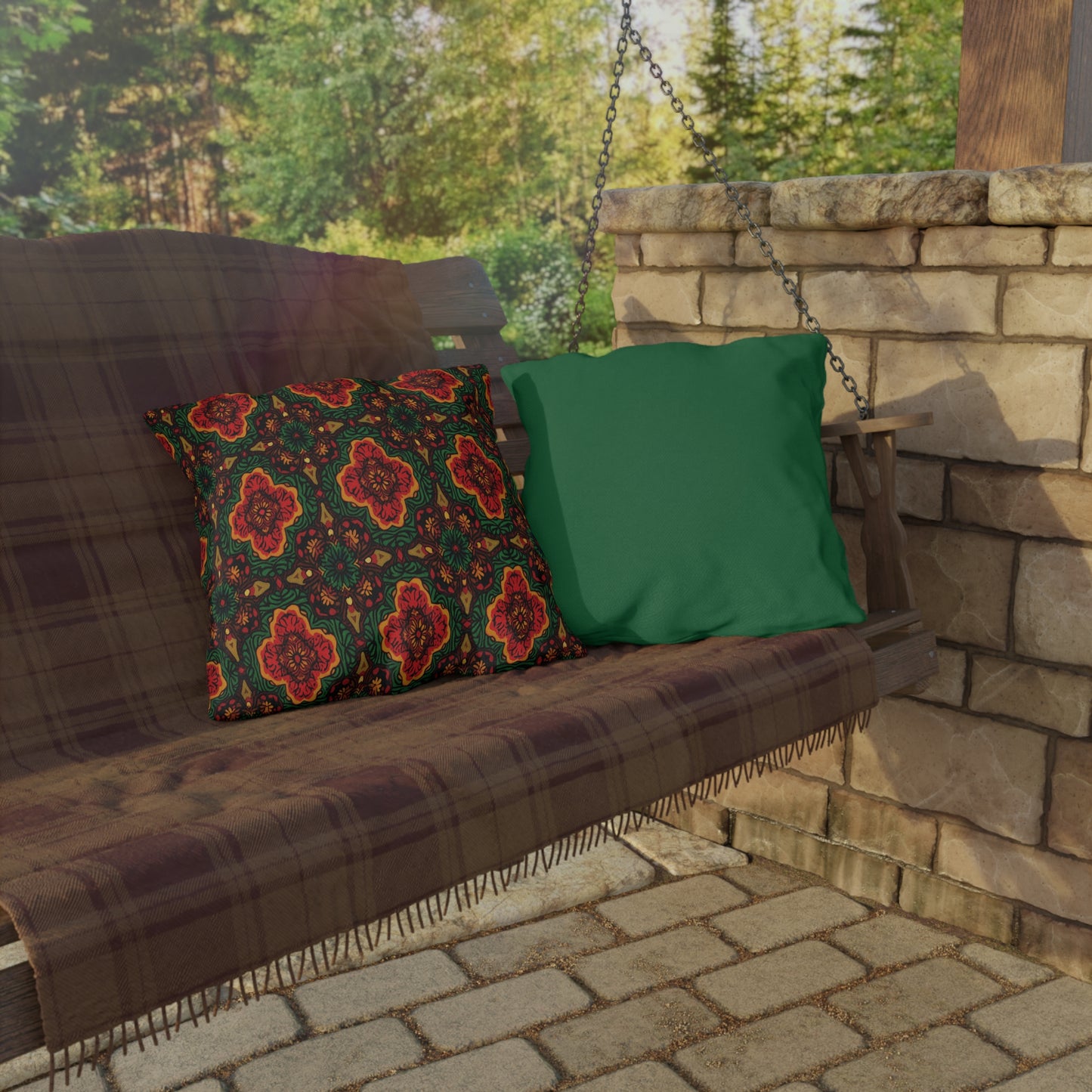 A wooden porch swing with a plaid blanket draped over it holds two pillows: one with an African-inspired red and green floral pattern, and one solid green. The swing hangs near a stone wall with trees and sunlight in the background.