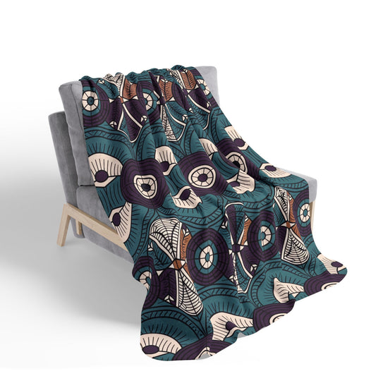 A modern gray armchair draped with a sherpa fleece blanket featuring a bold, abstract pattern in teal, navy, beige, and brown. The geometric and circular motifs create an eye-catching contrast that elevates cozy home decor.