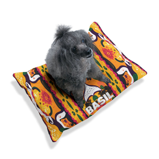 A small gray poodle sits on a colorful rectangular dog bed featuring bold African-inspired orange, yellow, and green patterns with pizza and basil leaves. The word “BASIL” is printed in large white letters near the bottom right corner of the bed.
