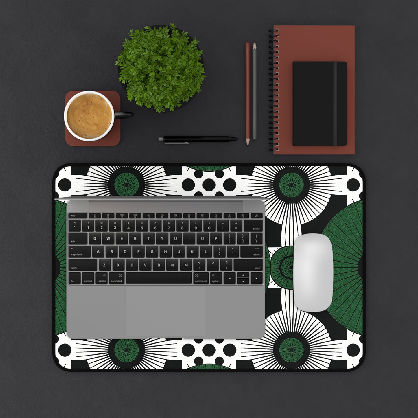 A laptop and white mouse rest on an ergonomic desk mat with black, white, and green patterns. Beside them sit a brown notebook, pencil, black pen, small potted plant, and a coffee cup—all arranged on a dark surface.