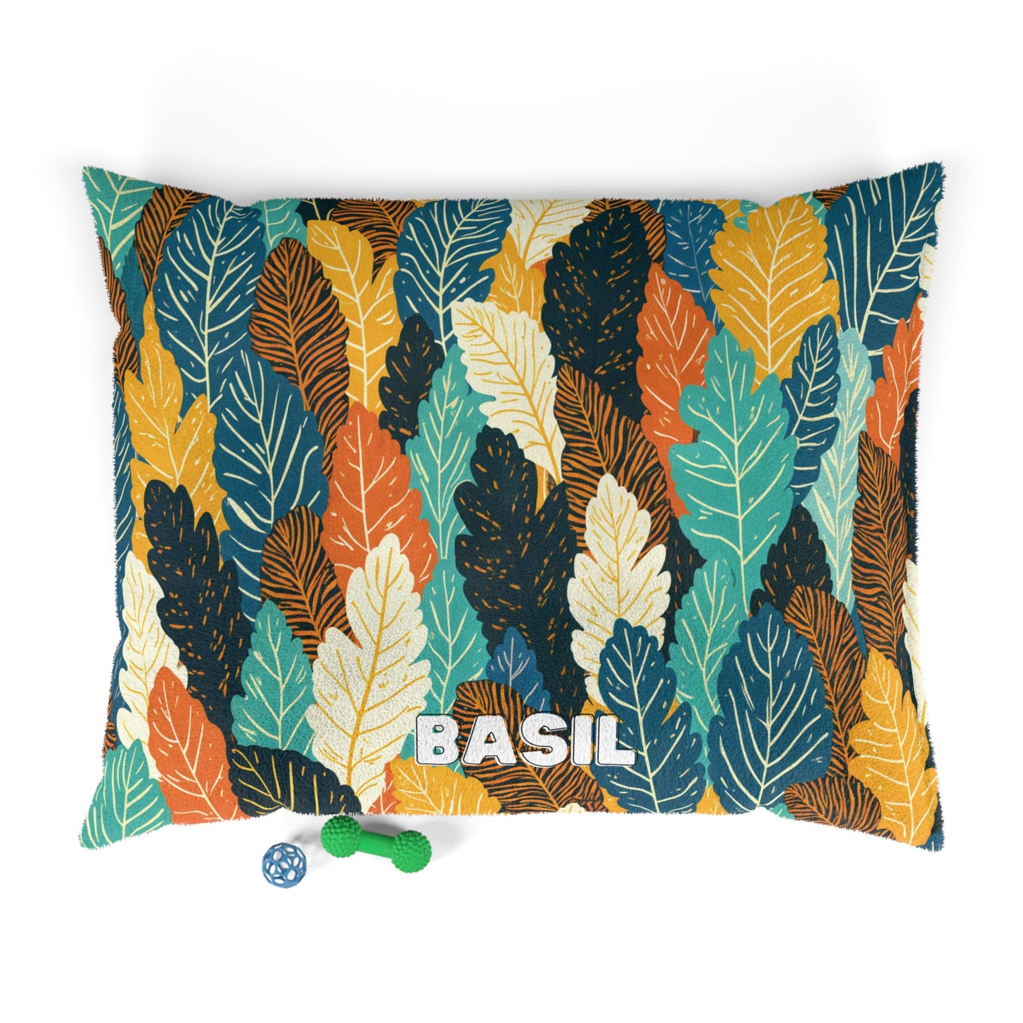 A rectangular pillow with a colorful leaf pattern in teal, yellow, orange, brown, and cream offers luxurious comfort. The word BASIL is printed in white letters. Two dog toys—a green textured bone and a blue ball—are placed nearby on a white background.