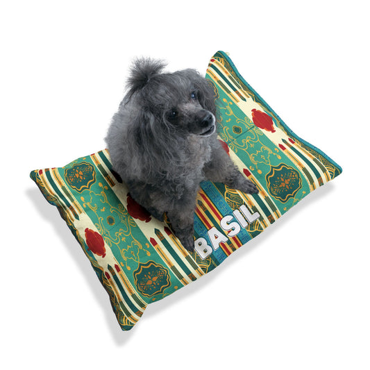 A small, fluffy gray poodle sits on a colorful pillow with a teal, yellow, and red geometric pattern. The name BASIL is printed in large white letters on this personalized pet bed. The background is plain white.