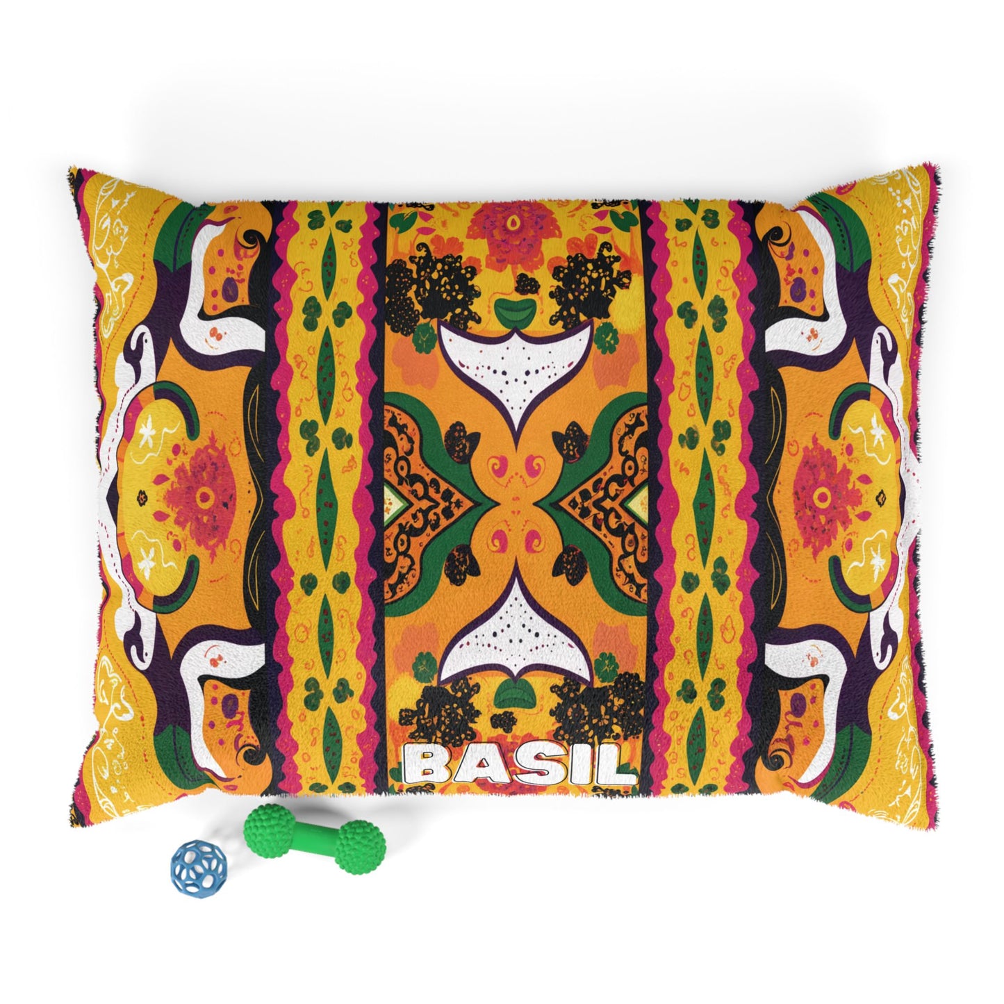 A decorative rectangular pillow with an African-inspired, colorful, symmetrical pattern in orange, green, yellow, black, and white features floral and abstract shapes. The word BASIL appears near the bottom. Two dog toys are nearby: a blue ball and a green bone.