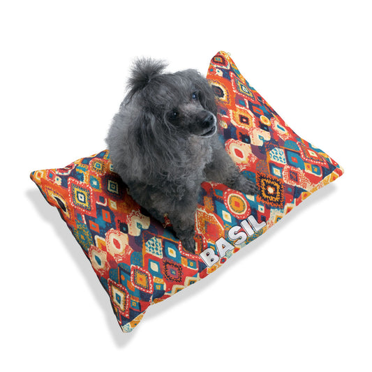 A small gray poodle with curly fur sits on a colorful African-inspired dog bed featuring geometric shapes in red, blue, yellow, and orange. The word BASIL is printed in bold white letters on the bed. The background is plain white.