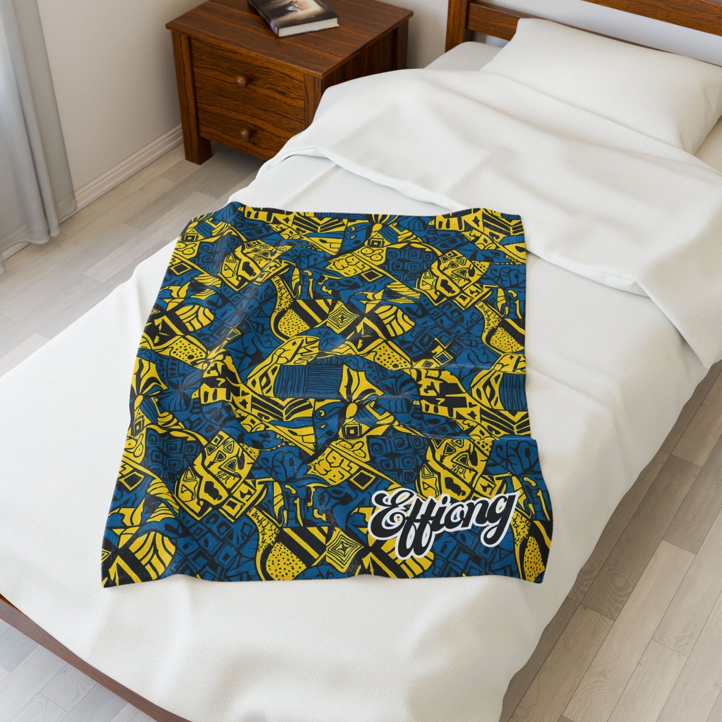 A colorful African-inspired blanket with bold blue and yellow geometric patterns and the word Effiong in white script is spread over a neatly made bed beside a wooden nightstand in a bright, minimalist bedroom.