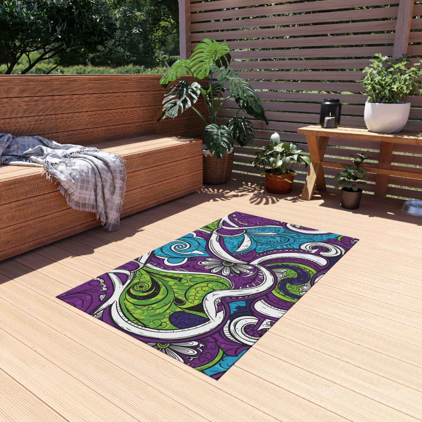 A colorful rectangular polyester chenille rug with swirling abstract patterns in purple, green, blue, and white lies on a light wooden deck. Potted plants and a bench add comfort to this cozy outdoor space featuring weather-resistant rugs.