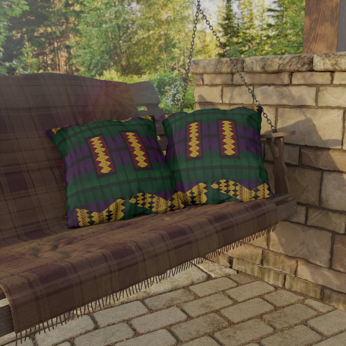A wooden porch swing with a plaid blanket and two colorful outdoor pillows sits on a stone patio. The pillows feature green, purple, and gold geometric designs crafted from UV-resistant fabric. Sunlight filters through trees in the background, casting soft shadows.