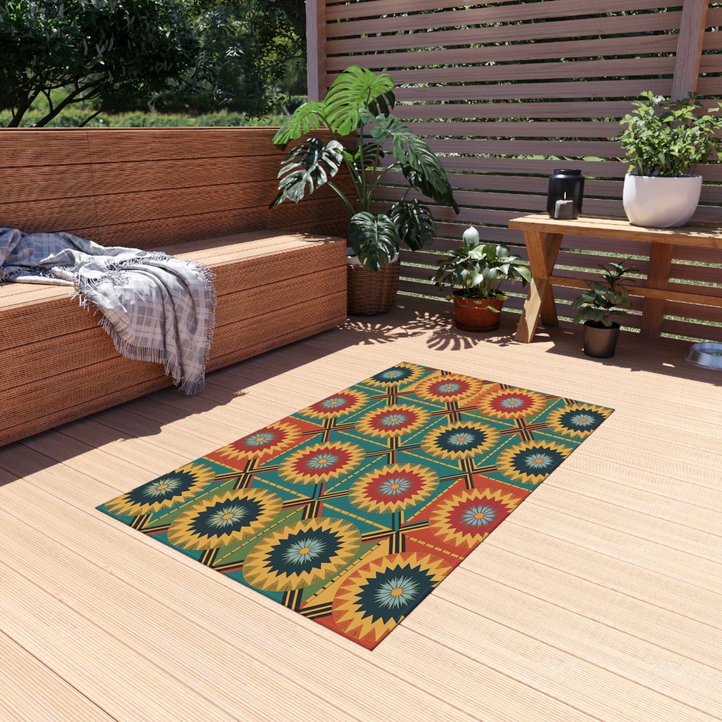 A colorful African-inspired outdoor rug with red, yellow, and green starburst patterns lays on a light wooden deck. Its no-slip backing ensures safety, while wicker furniture and potted plants create a cozy, modern outdoor seating area.