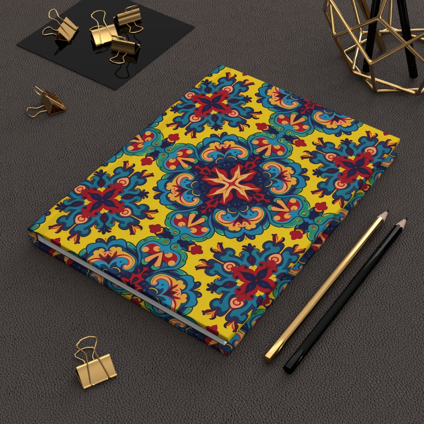 A colorful hardcover journal with an ornate, symmetrical floral pattern in blue, red, and yellow lies on a textured brown surface. Nearby are a gold pen, black pencil, gold binder clips, and a geometric gold wire decor piece.