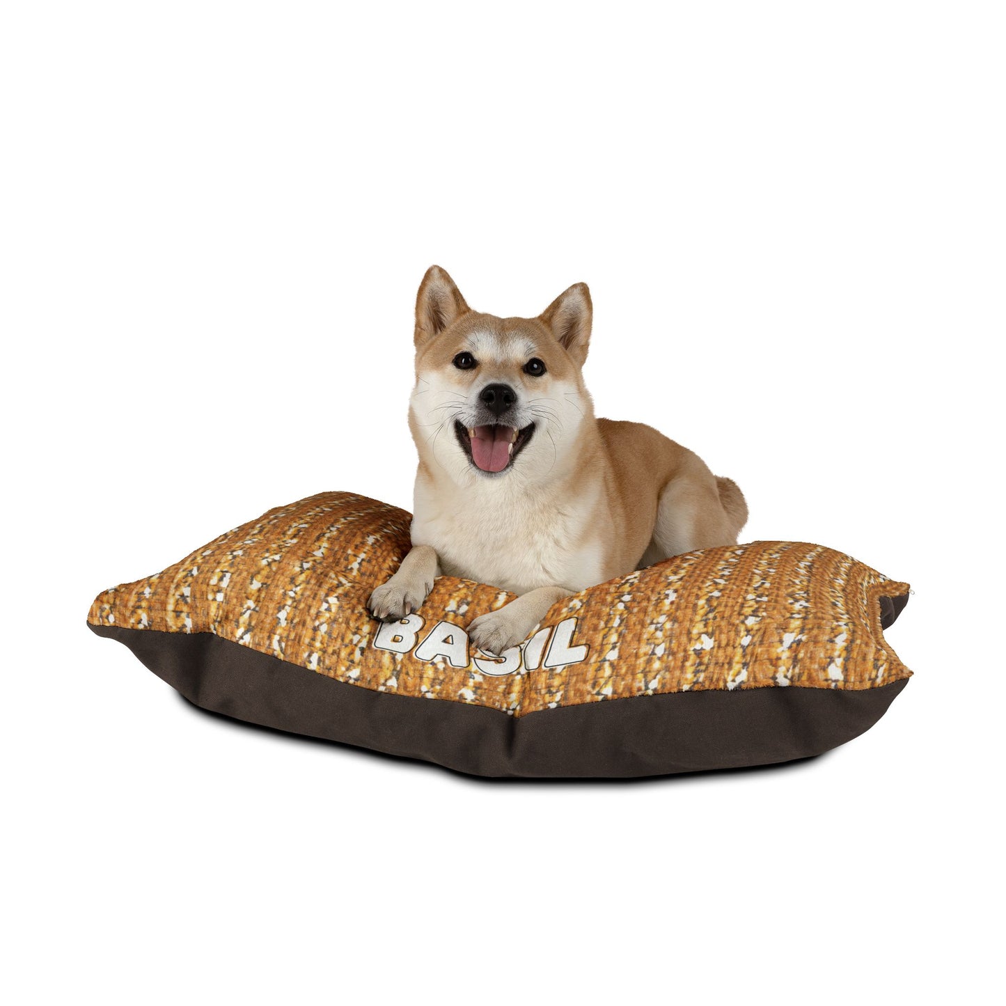 A happy Shiba Inu dog with tan fur lies on a stylish pet bed with a brown and orange pattern. The cozy pet pillow features the name BASIL printed in white letters. The background is plain white.