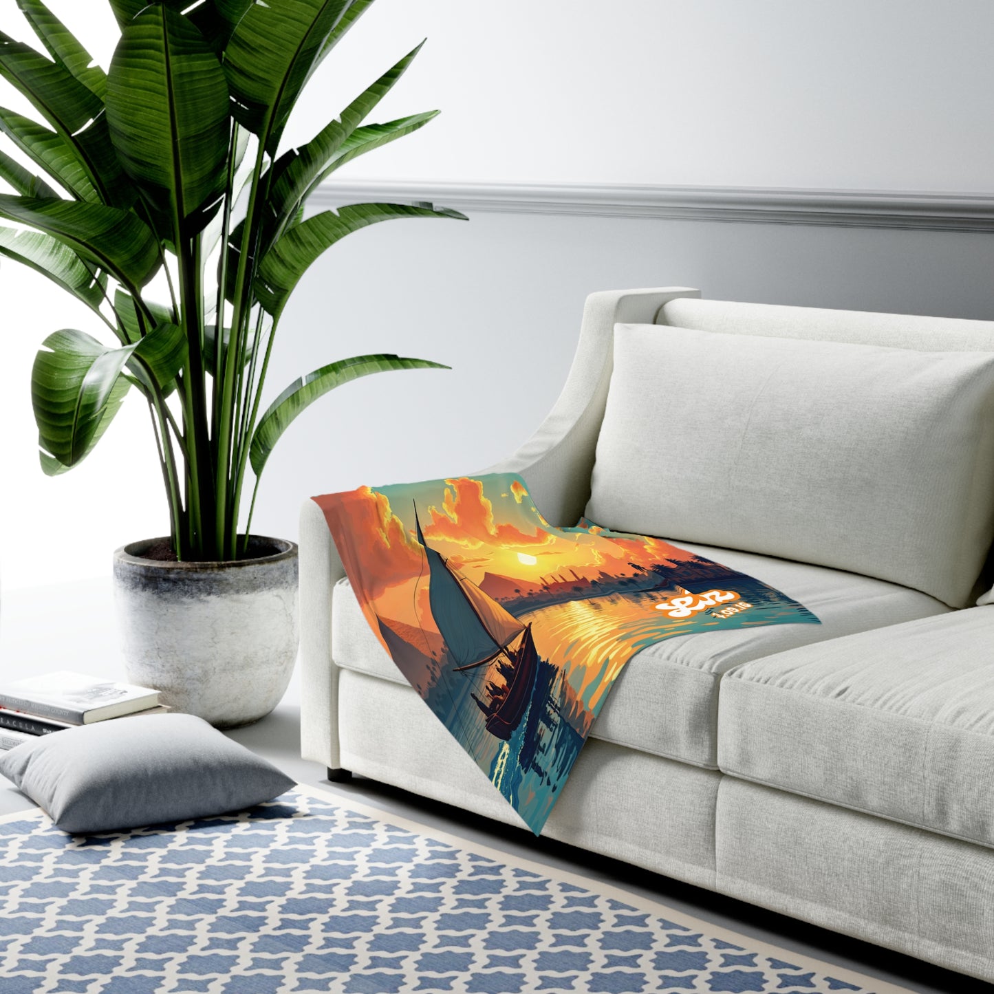 A white sofa with a colorful custom swaddle blanket draped over the side, depicting a sailboat at sunset. A large potted plant stands nearby, and a blue and white patterned rug with a cushion is on the floor in this modern living room setting.