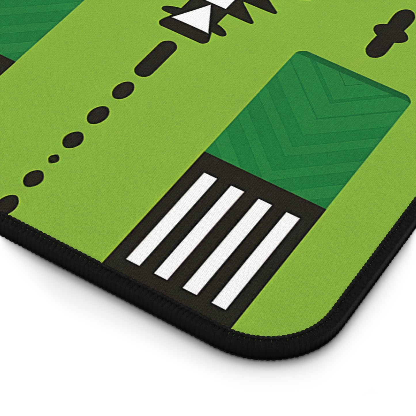 A close-up of a green neoprene desk mat with geometric patterns, including black dots, white stripes, green chevrons, and abstract shapes. The design is bordered by a black edge and appears smooth and modern.