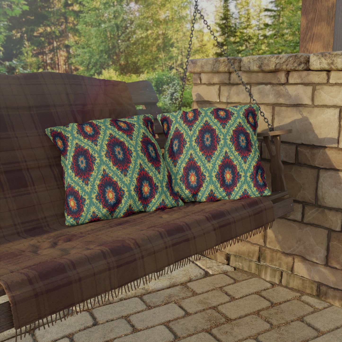 A wooden porch swing with a brown plaid blanket and two African-inspired, colorful patterned cushions sits on a stone patio next to a stone pillar. Lush green trees are visible in the background, with sunlight casting soft shadows.