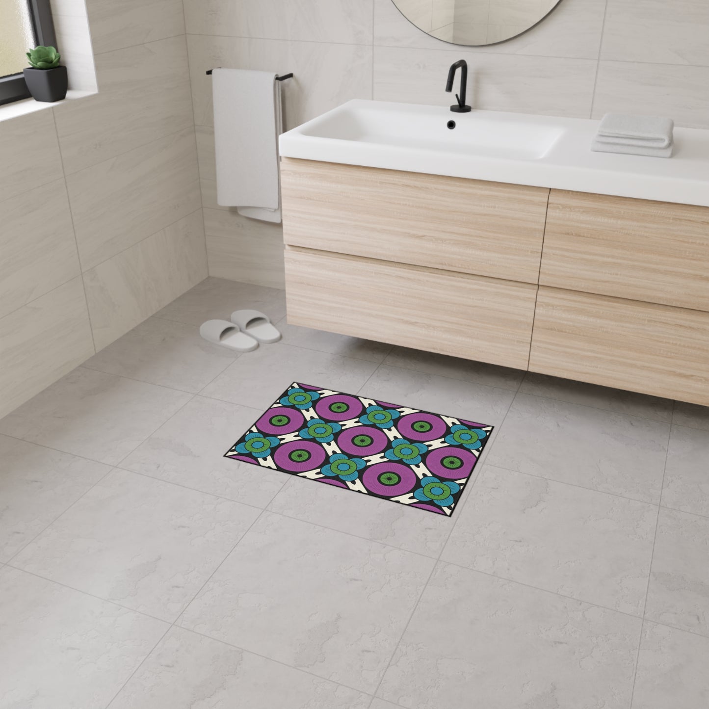 A modern bathroom with a light wood vanity, white countertop, circular mirror, towel, white slippers, and a colorful custom floor mat featuring green, purple, and blue circular patterns on the light gray tiled floor.