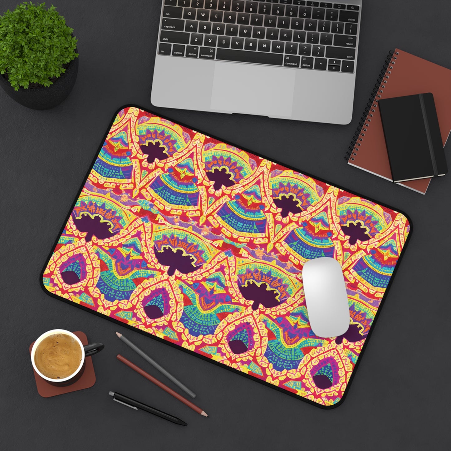 A colorful workspace accessory, this vibrant neoprene desk mat features an abstract pattern in red, yellow, blue, and purple. It brightens a dark desk beside a laptop, notebooks, potted plant, coffee cup, two pencils, and a white mouse.