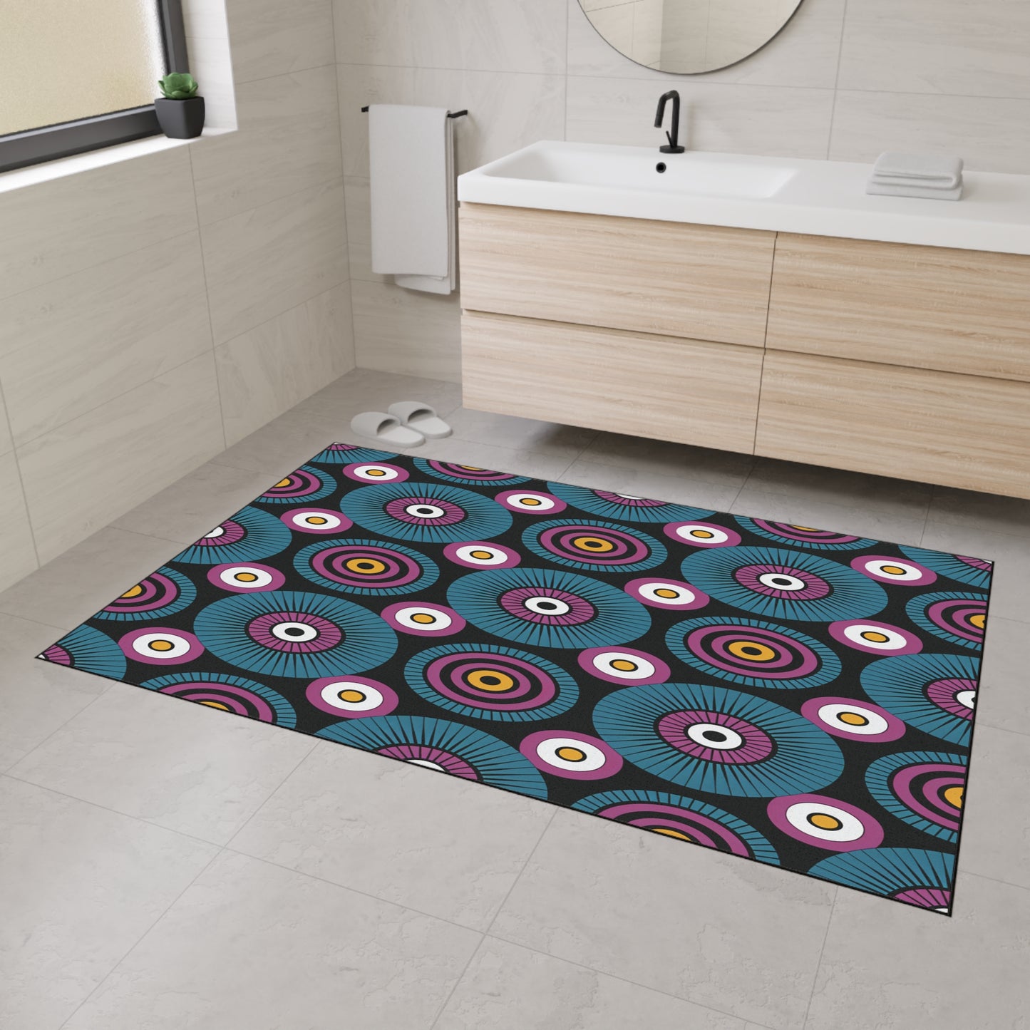 A modern bathroom with light beige tiled floors and walls features a large, colorful bath mat with concentric circle patterns and non-slip rubber backing, adding a touch of African-inspired aesthetics beside the wood vanity, round mirror, towel, and window.