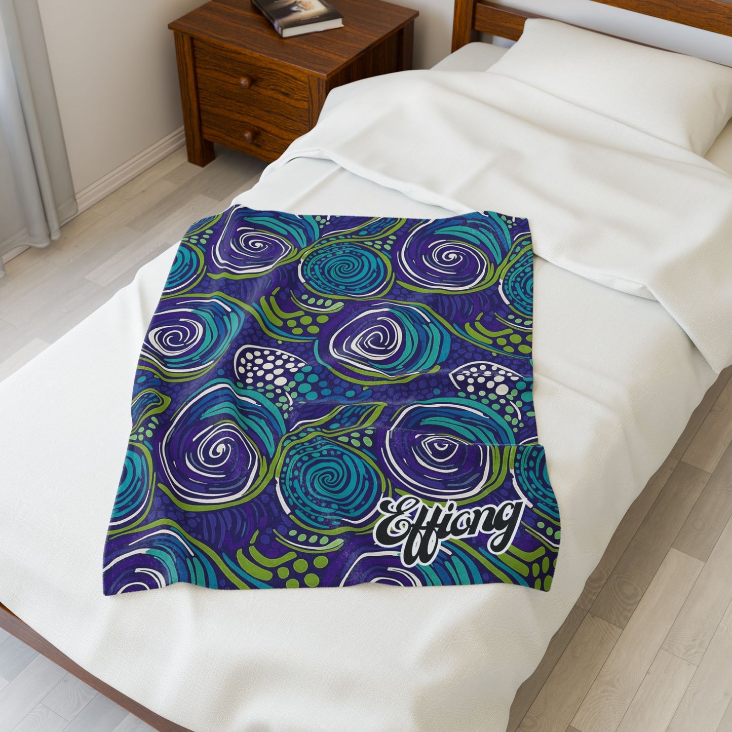 A colorful personalized velveteen blanket with swirling abstract patterns in blue, green, purple, and white lies on a neatly made bed. The word Effiong is printed in cursive on the corner. A wooden nightstand with a book sits beside the bed.