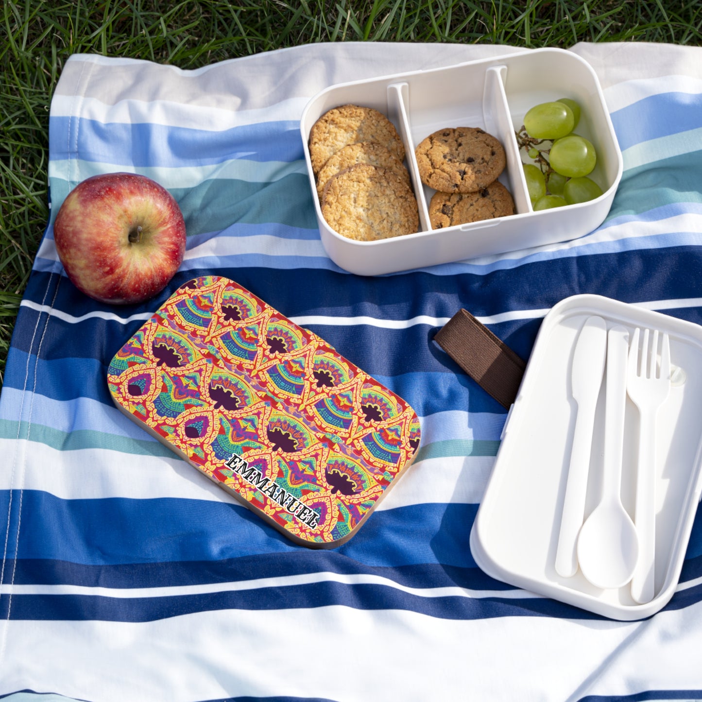A colorful, personalized lunch box labeled IMMANUEL sits on a blue-striped blanket on grass. The BPA-free lunchbox is open to show cookies and green grapes, with white cutlery in an empty container and a red apple nearby.