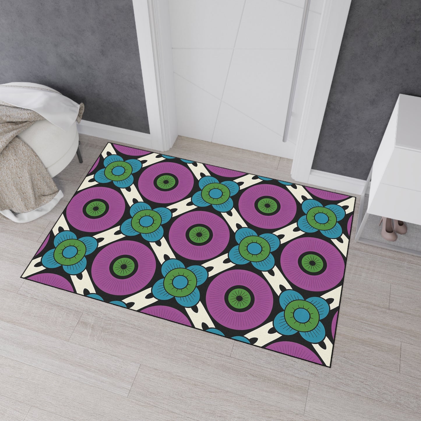 A colorful geometric rug with a bold pattern of large purple circles, teal and blue floral shapes, and black accents features non-slip rubber backing. It lies on a light wood floor in front of a white door, with a beige blanket and white furniture nearby.
