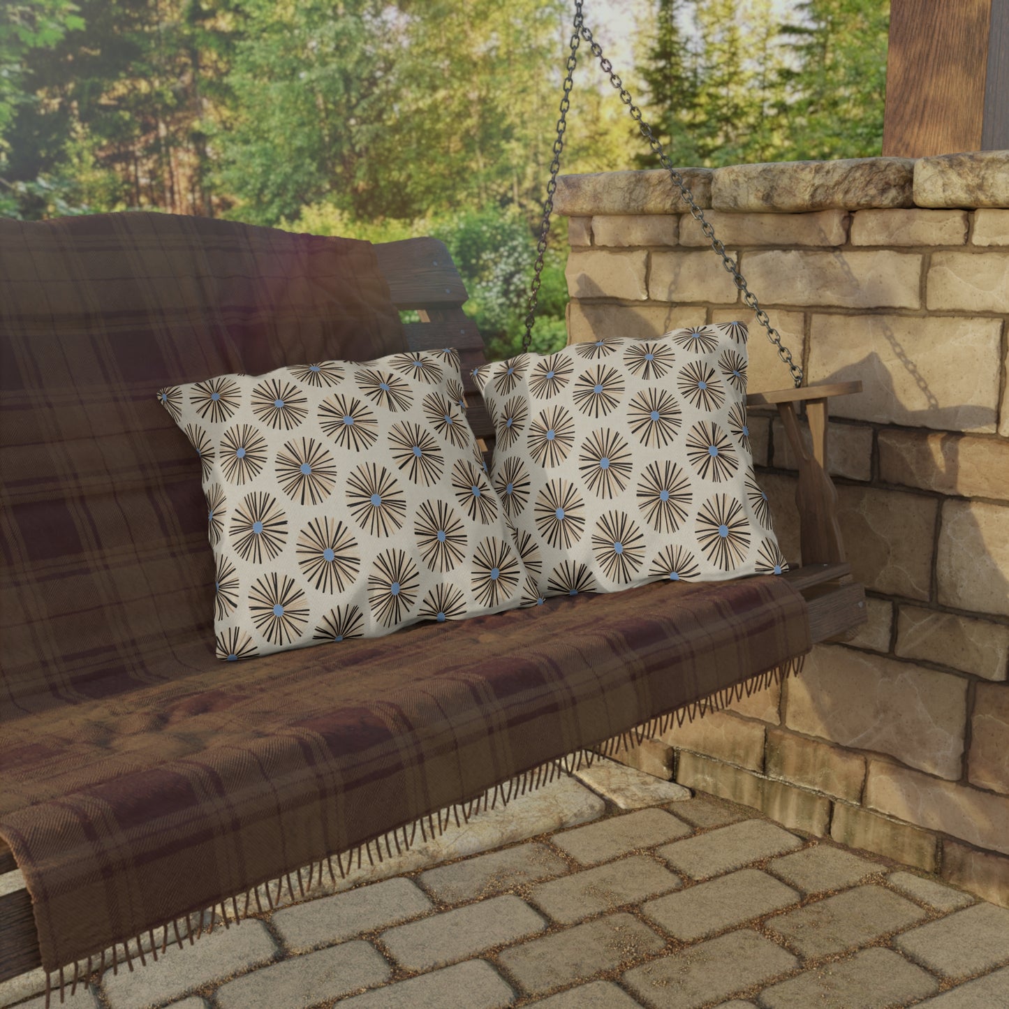 A wooden porch swing covered with a brown plaid blanket holds two African-inspired patterned cushions. The swing hangs by metal chains near a stone wall, with a paved stone floor beneath and green trees glowing in the sunlight behind.