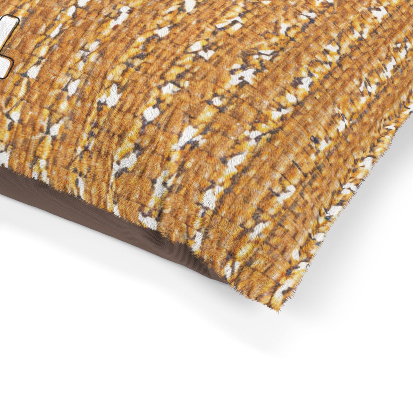 A close-up of a rectangular, stylish pet bed pillow with a textured, woven golden-brown pattern and white highlights on top, plus solid dark brown fabric on the side. The pillow’s corner is visible against a plain white background.