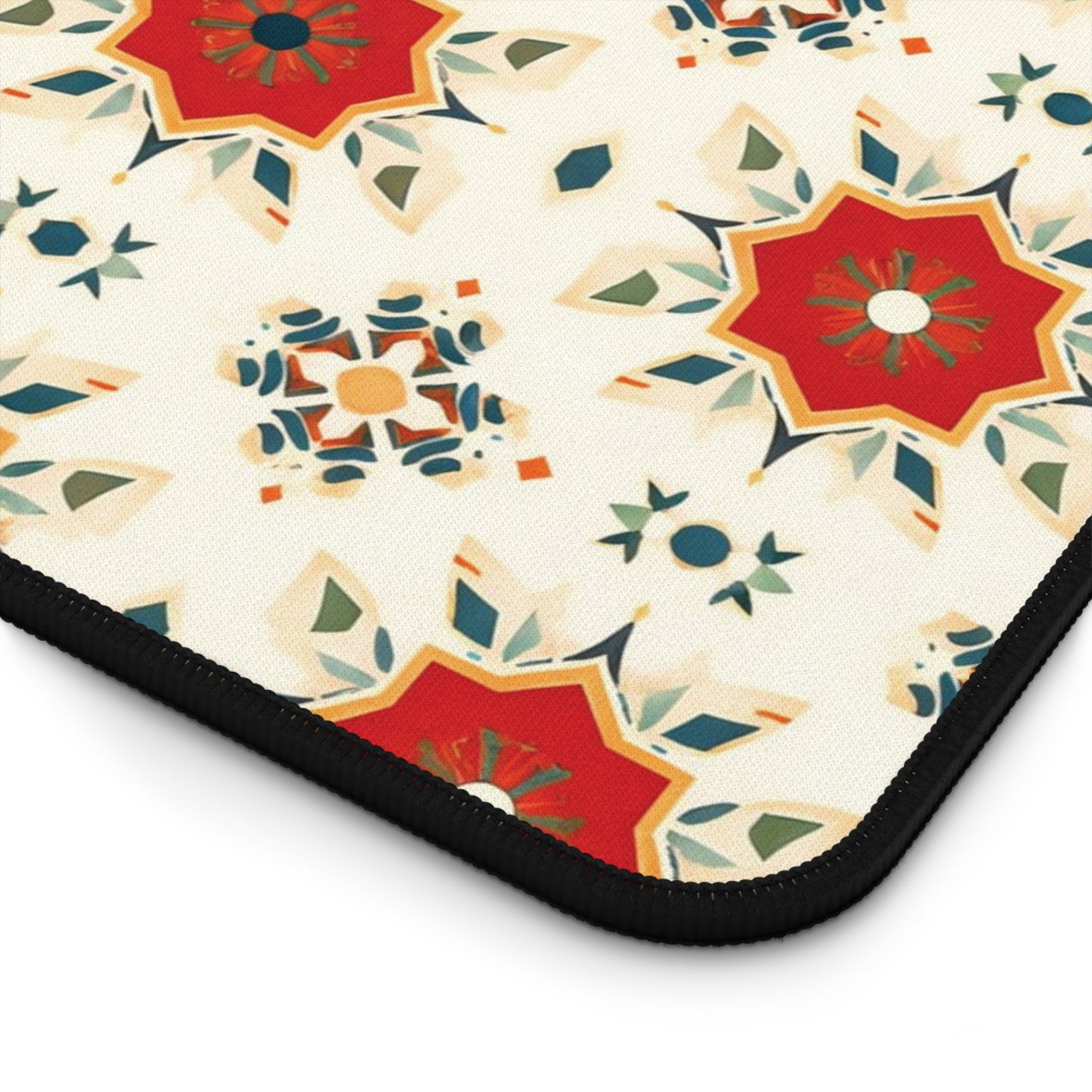 A close-up of a neoprene desk mat displays bold, geometric floral designs in red, green, blue, and orange on a cream background. The motifs have a starburst effect, and the mat has a black, rounded border on one edge—a truly colorful workspace accessory.