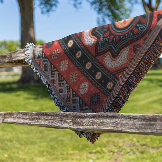 An African-inspired, fringed blanket with red, green, and cream geometric designs is draped over a weathered wooden fence outdoors. The background features green grass, leafy trees, and a clear blue sky on a sunny day.