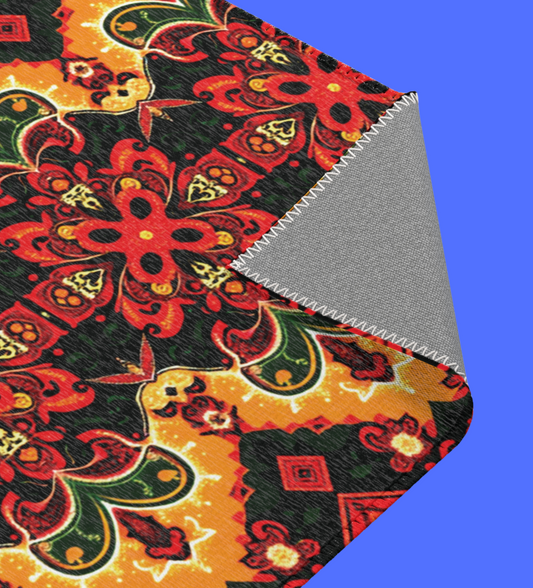 A blanket with a vibrant red, yellow, green, and black mandala-like floral pattern inspired by African design rugs is shown on a blue background. The corner is folded over, revealing a plain gray underside with visible zigzag stitches along the edge.