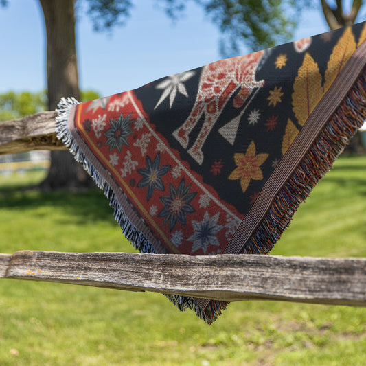 A vibrant throw blanket with fringed edges, featuring stars, leaves, and an animal in a cozy weave, is draped over a rustic wooden fence outdoors. Green grass, trees, and blue sky create a blurred backdrop.