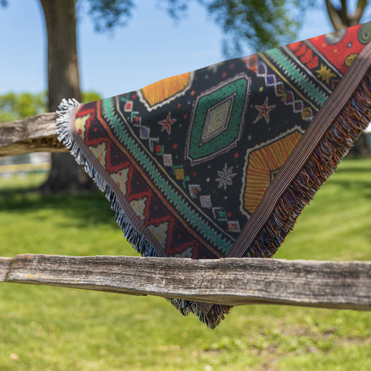 A colorful, African-inspired woven blanket with geometric and star patterns hangs over a rustic wooden fence outdoors, framed by green grass, trees, and a sunny sky. The fringed edges showcase vibrant reds, greens, yellows, and blacks.