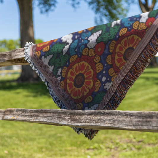A colorful, African-inspired woven blanket with a fringed edge, featuring large floral patterns in red, yellow, and green, is draped over a rustic wooden fence. The scene includes bright green grass and leafy trees beneath a clear blue sky.