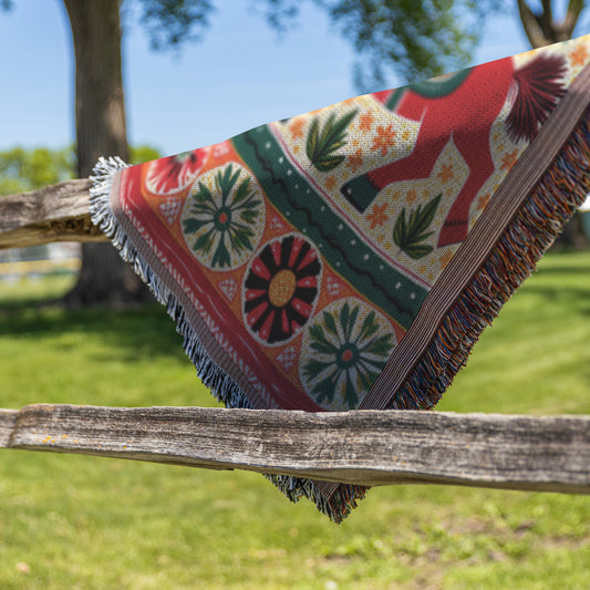A colorful, African-inspired woven blanket with floral and geometric patterns and a red horse design hangs draped over a wooden fence in a grassy area with green trees and a blue sky in the background.