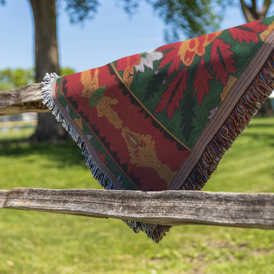 A colorful, African-inspired fringed blanket with red, green, and yellow floral patterns is draped over a rustic wooden fence. Green grass, leafy trees, and a blue sky in the background suggest a sunny outdoor setting.