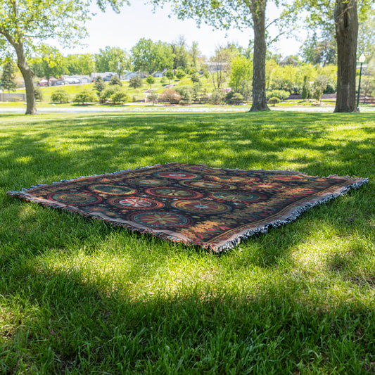 A vibrant throw blanket with fringed edges is spread out on green grass in a sunny park. Trees cast dappled shadows, and landscaped shrubs, a pond, and a blue sky appear in the background, suggesting a peaceful outdoor setting.