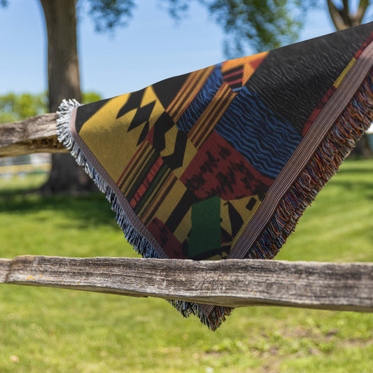 A vibrant throw blanket with colorful geometric patterns and fringed edges is draped over a rustic wooden fence outdoors. The scene features green grass, leafy trees, and a clear blue sky, creating a sunny and peaceful atmosphere.