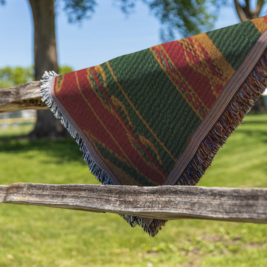 A colorful African-inspired woven blanket with green, red, and yellow patterns drapes over a rustic wooden fence. The fringed edges and vibrant design stand out against lush grass and leafy trees, evoking a peaceful outdoor setting.
