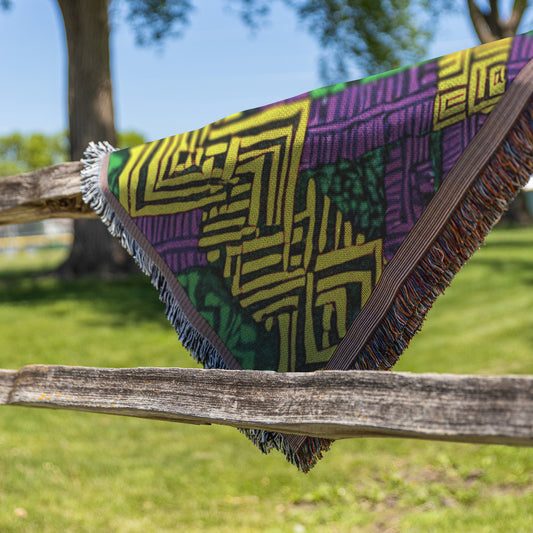 A vibrant throw blanket with a geometric West African print and fringed edges is draped over a rustic wooden fence outdoors. The blanket’s bright green, yellow, and purple designs stand out against lush grass and leafy trees under a blue sky.