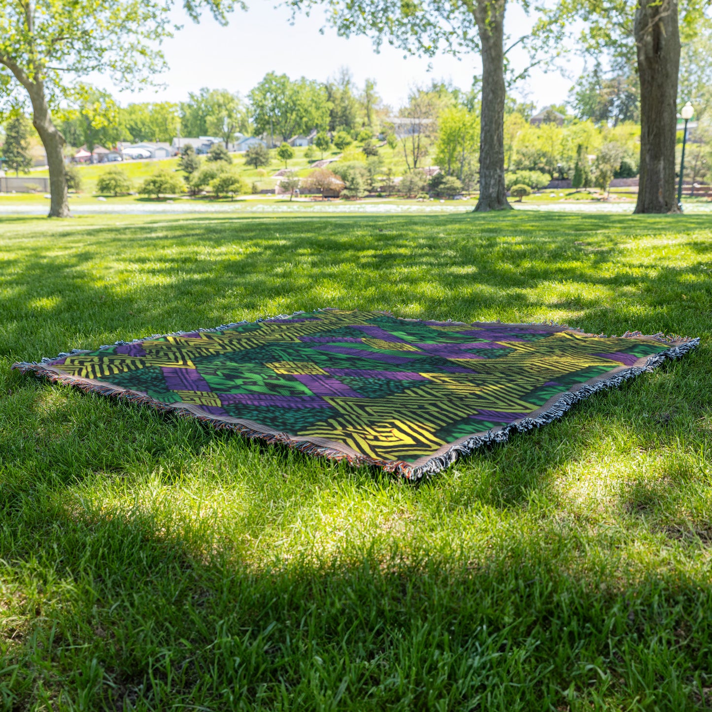 A vibrant throw blanket with green, purple, and yellow geometric patterns lies on green grass in a sunlit park. Trees cast dappled shade, with more greenery and houses visible in the background under a clear sky.