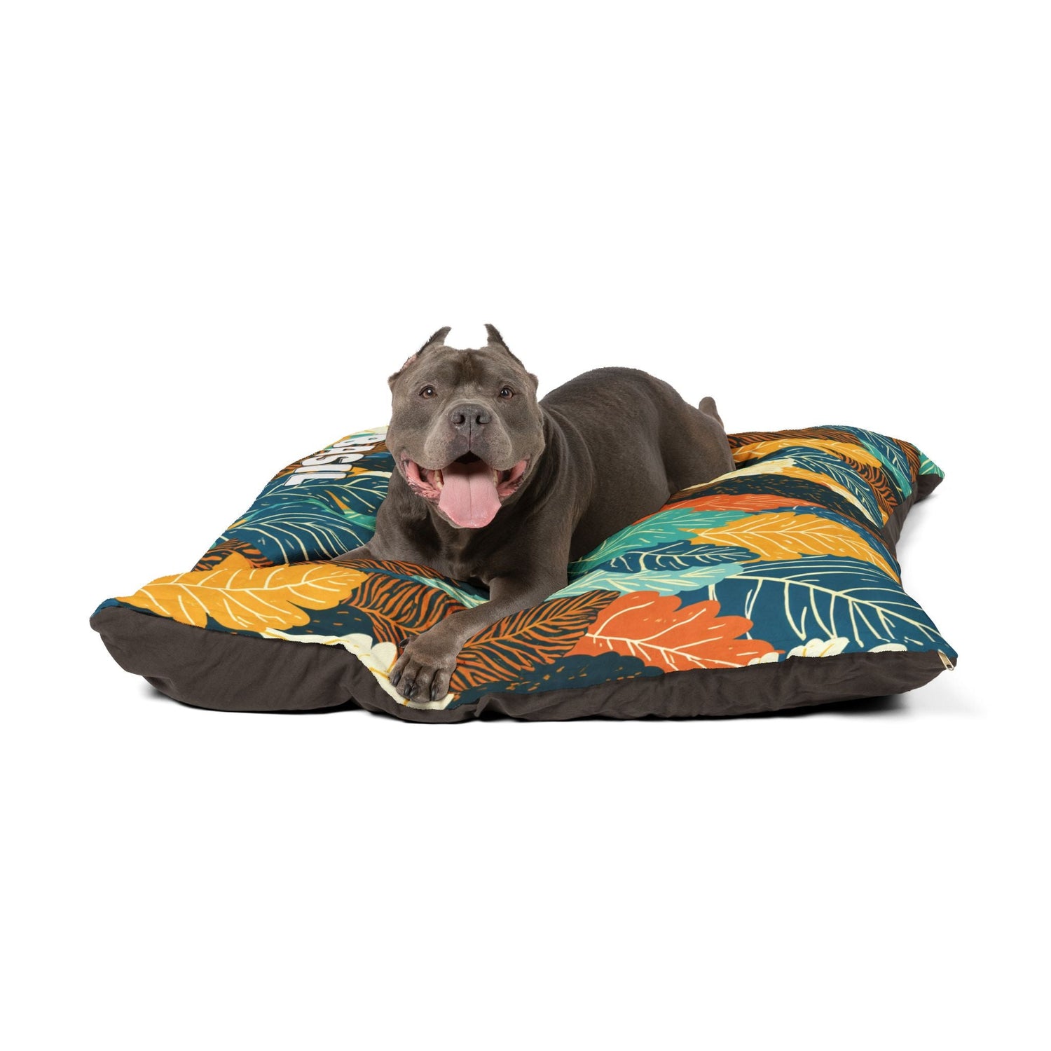 A happy gray pit bull with cropped ears lies on a colorful, leaf-patterned dog bed, panting with its tongue out against a white background.
