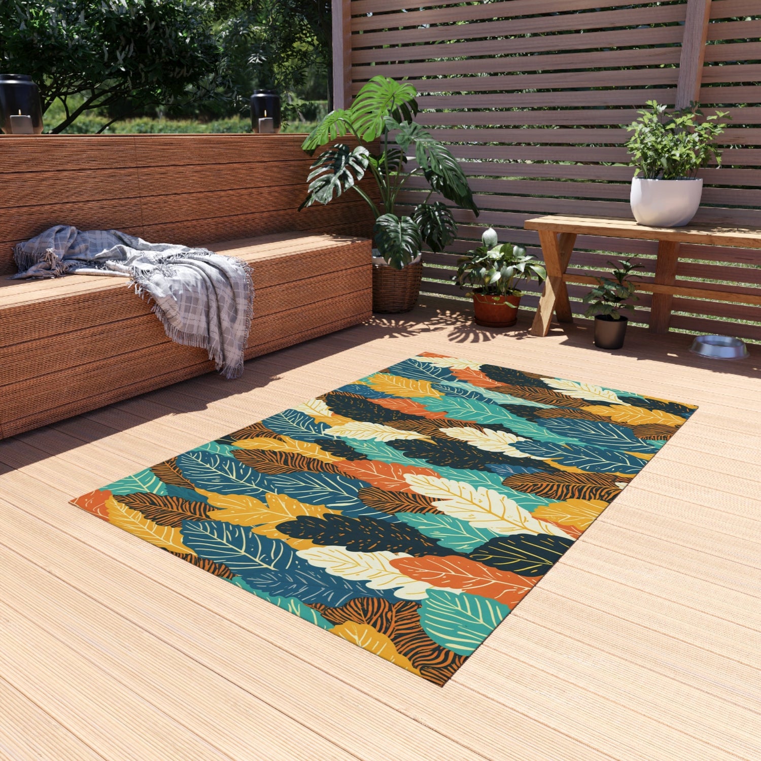 A colorful leaf-patterned rug lies on a light wooden patio beside a wicker bench with a gray throw, green plants, a wooden table, and potted plants, under bright natural light.