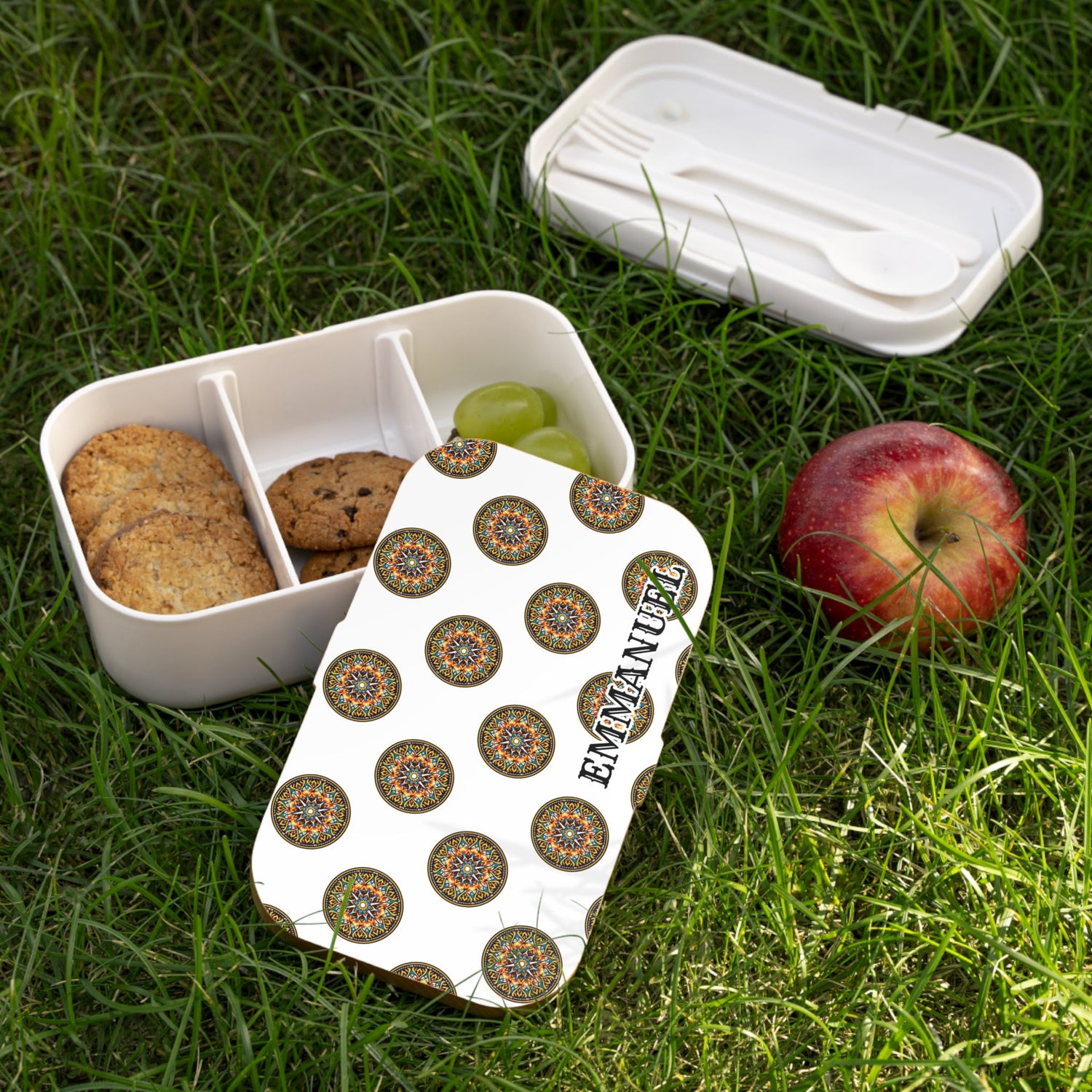 A bento box with cookies and grapes sits open on grass, its lid decorated with circular patterns and the name “Emmanuel.” An apple and a spoon and fork rest beside the box.