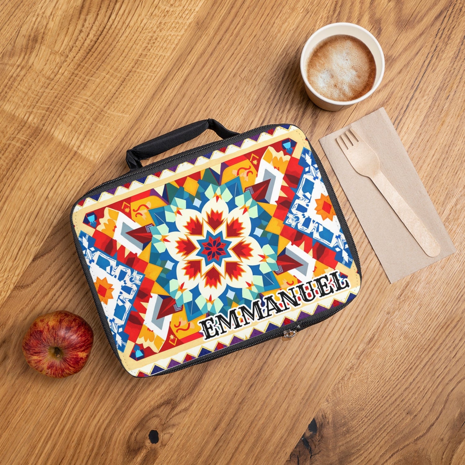 A colorful, patterned lunchbox labeled “EMMANUEL” sits on a wooden table near a coffee cup, wooden fork and knife on a napkin, and a red apple.