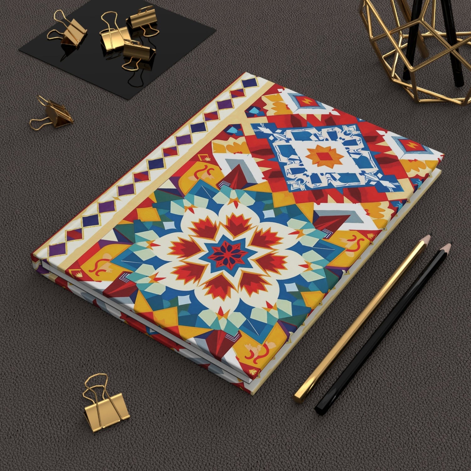 A journal with a colorful geometric mandala cover lies on a dark textured desk, surrounded by gold binder clips, a black pencil, a gold pen, and a decorative gold wire ornament.